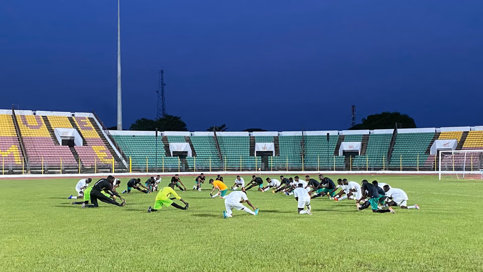 Two dozen players kneeling to stretch in a long row across a stadium pitch at dusk, the stands behind them empty